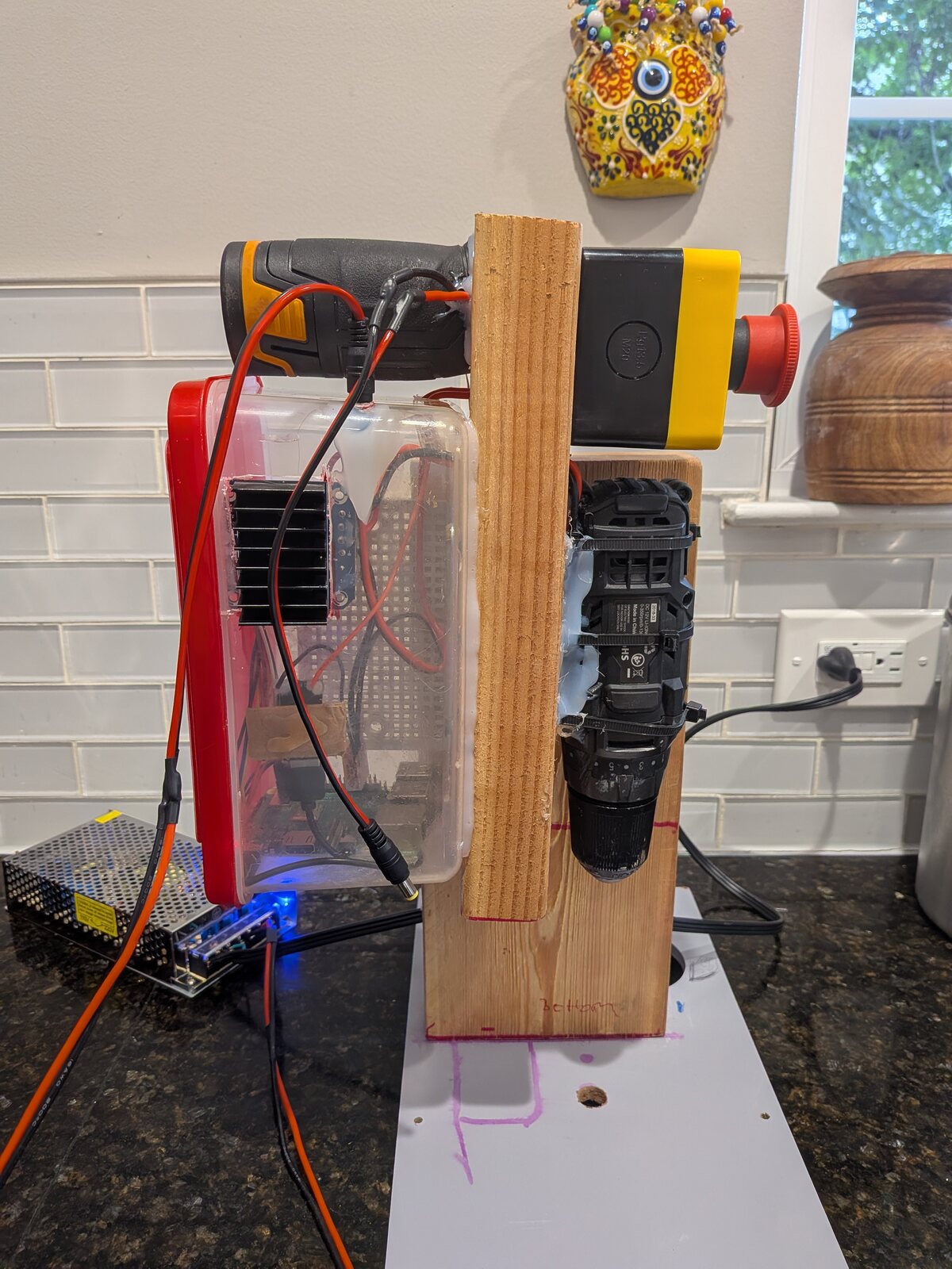 The rig on a kitchen counter, showing the drill in its clamp, the wooden board, the clear box of electronics and a Raspberry Pi at the base.