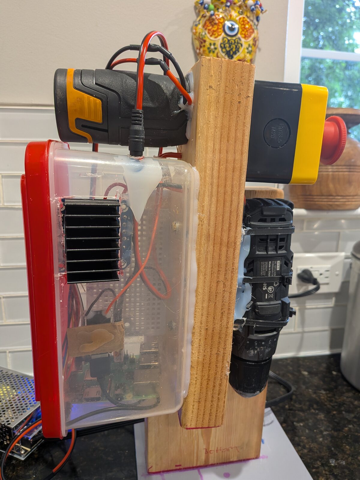 The rig standing on a kitchen counter. A drill clamped to a wooden board, a clear plastic box of electronics, and a yellow emergency stop button.